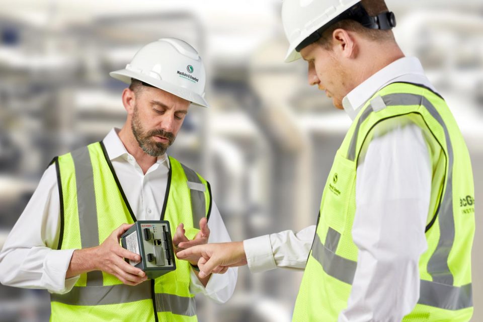 Two men wearing hard hats and yellow vests looking at a piece of instrumentation equipment